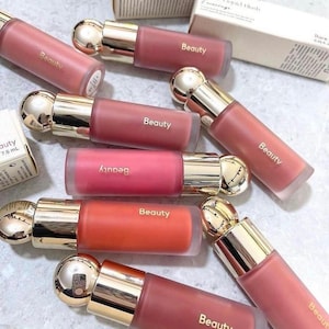 Puede incluir: Una variedad de botellas de colorete líquido en varios tonos de rosa y coral. Cada botella tiene una tapa dorada y la palabra "Beauty" impresa en el lateral. Las botellas están dispuestas sobre una superficie clara.