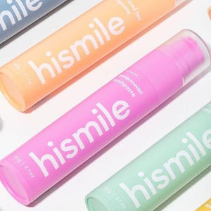 Puede incluir: Varios tubos de pasta de dientes de colores pastel, con la marca "hismile" en blanco. Los colores incluyen rosa, naranja y azul. Cada tubo indica un sabor y contiene 60g de pasta de dientes, con un tapón transparente.