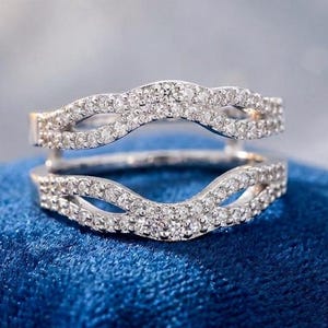 May include: A silver-toned ring guard featuring two curved bands connected at the sides. Each band is encrusted with small, clear, sparkling stones arranged in a pavé setting, creating a continuous, shimmering, wave-like design across the metal.