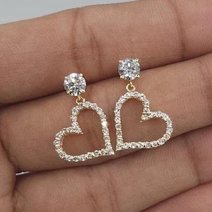 Può includere: Un paio di orecchini dorati con un design a forma di cuore. Ogni orecchino ha una gemma rotonda e trasparente nella parte superiore ed è delineato da pietre più piccole e scintillanti. Gli orecchini sono tenuti in una mano.