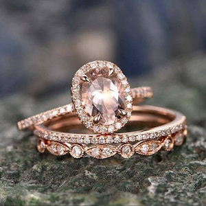 May include: Rose gold engagement ring set featuring an oval-cut peach-colored gemstone surrounded by a halo of small diamonds. The set includes a matching band and a delicate, diamond-studded band. The rings are displayed on a textured, green surface.