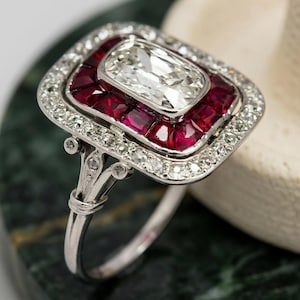 Könnte beinhalten: Ein Platinring mit einem zentralen rechteckigen Diamanten im Kissenschliff, umgeben von einem Kranz aus quadratisch geschliffenen, tiefroten Rubinen. Ein äußerer Rand aus kleinen runden Diamanten rahmt die Rubine ein, ergänzt durch kleine Diamanten.