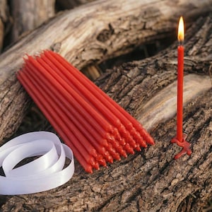 Pode incluir: Uma coleção de velas vermelhas cônicas, uma vela acesa e uma fita branca em uma superfície de madeira texturizada. A vela acesa tem uma pequena base vermelha. As velas apagadas estão agrupadas.