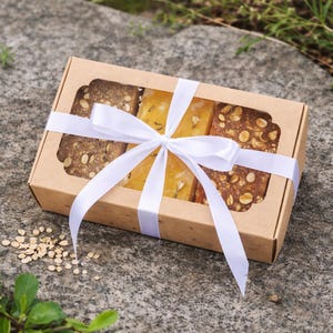 Puede incluir: Una caja de regalo de cartón marrón con una ventana transparente, que contiene varias barras de jabón. Los jabones son de varios colores y texturas, y la caja está atada con una cinta blanca. Presentado sobre una superficie de piedra.
