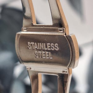 May include: Close-up of a stainless steel watch clasp. The clasp is silver with the words "STAINLESS STEEL" embossed on the surface. The watch band is visible on either side of the clasp.