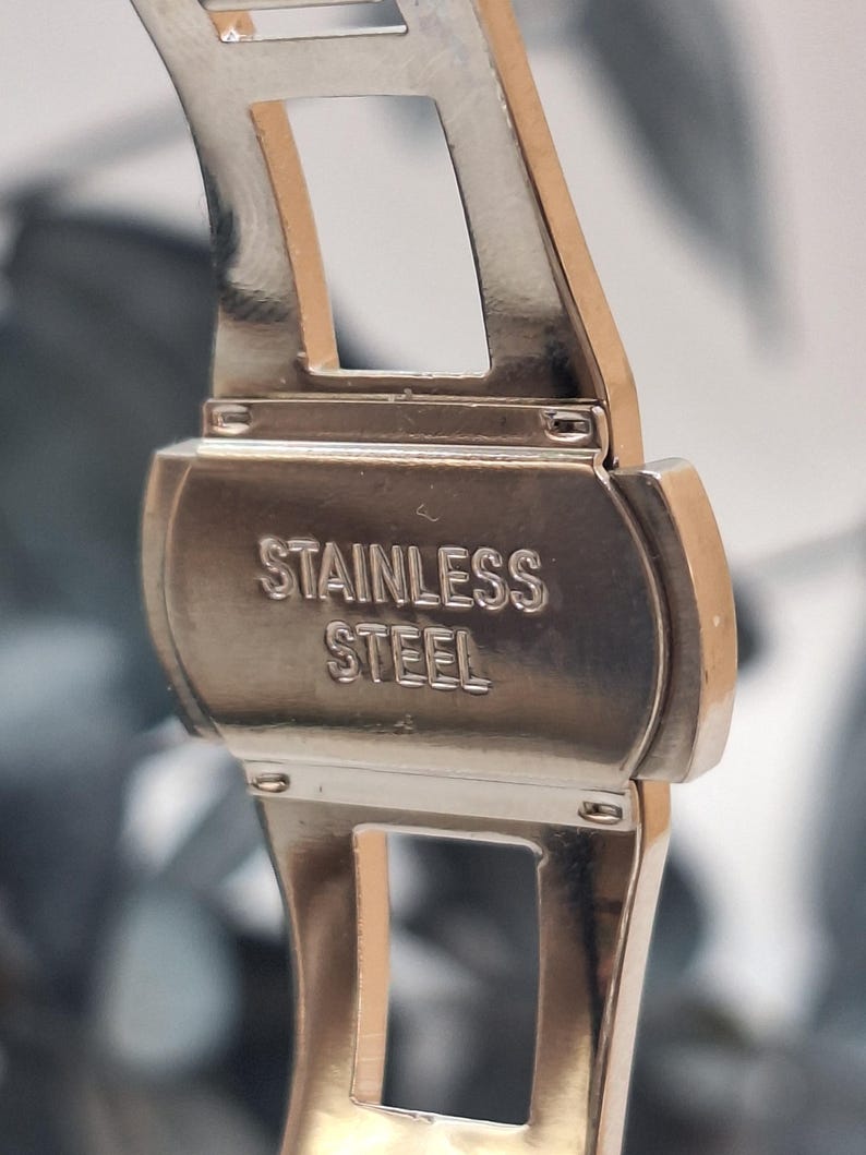 May include: Close-up of a stainless steel watch clasp. The clasp is silver with the words "STAINLESS STEEL" embossed on the surface. The watch band is visible on either side of the clasp.
