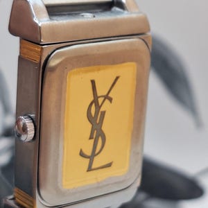 May include: Close-up of a rectangular wristwatch with a silver-tone metal band and case. The watch face is gold with a black logo. The watch has a silver-tone crown.
