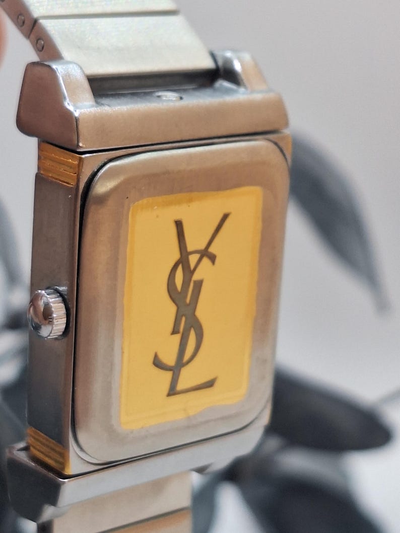 May include: Close-up of a rectangular wristwatch with a silver-tone metal band and case. The watch face is gold with a black logo. The watch has a silver-tone crown.