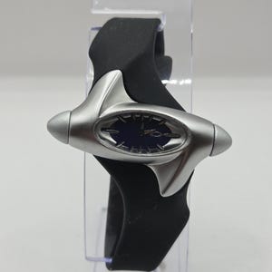 May include: A wristwatch featuring a dark blue oval dial with silver-tone hour markers and hands, encased in a smooth, matte silver-tone frame with pointed ends. The watch is attached to a flexible, solid black silicone strap that wraps around.