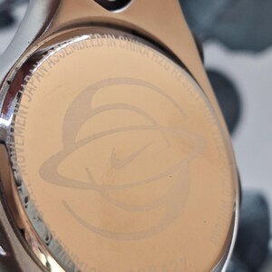 May include: Close-up of a stainless steel watch back with the Nike logo and text. The watch back is a light gold color with the Nike logo in the center. The text includes "MOVEMENT JAPAN ASSEMBLED IN CHINA R20 RESIST 30M".