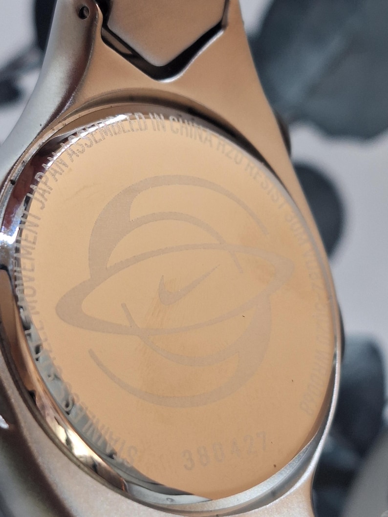 May include: Close-up of a stainless steel watch back with the Nike logo and text. The watch back is a light gold color with the Nike logo in the center. The text includes "MOVEMENT JAPAN ASSEMBLED IN CHINA R20 RESIST 30M".