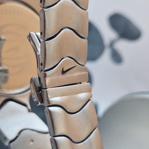 May include: Close-up of a silver-toned watch band with a repeating wave-like design. The Nike swoosh logo is visible on one of the links. The watch band is made of metal and has a polished finish. The watch face is partially visible.