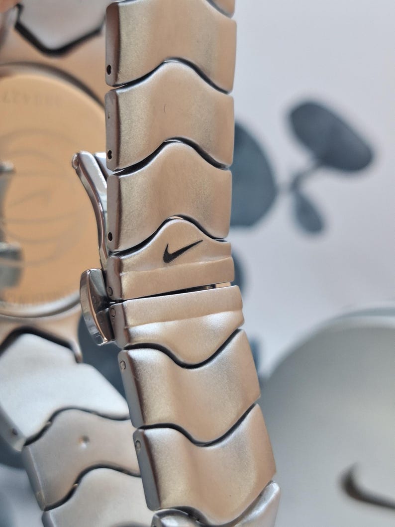 May include: Close-up of a silver-toned watch band with a repeating wave-like design. The Nike swoosh logo is visible on one of the links. The watch band is made of metal and has a polished finish. The watch face is partially visible.