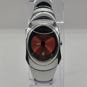 Könnte beinhalten: Eine Armbanduhr aus Edelstahl mit einem polierten, silberfarbenen Metallarmband und einem abgerundeten Gehäuse. Das Zifferblatt ist in einem tiefen Metallic-Rot gehalten, hat einen schwarzen Rand, silberne Zeiger und ein Datumsfenster mit der 3.