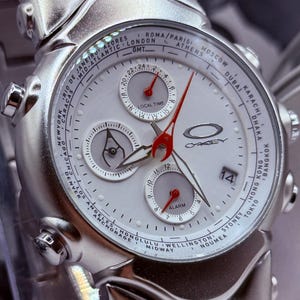 Könnte beinhalten: Silberfarbene Oakley Armbanduhr mit weißem Zifferblatt, mehreren Hilfszifferblättern und roten Zeigern. Die Uhr zeigt Weltzeitzonen und das Datum an. Das Armband ist ebenfalls silberfarben.