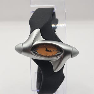 May include: A wristwatch featuring a matte silver-toned metal casing with an elongated, oval-shaped face and a light orange dial. The watch is attached to a smooth, black silicone strap that wraps around the central metallic housing.