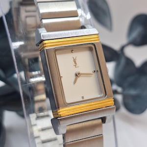May include: A silver-toned wristwatch with a rectangular face and a gold-colored border. The watch features a white dial with gold hands and a logo. The watch band is made of silver-toned metal links.