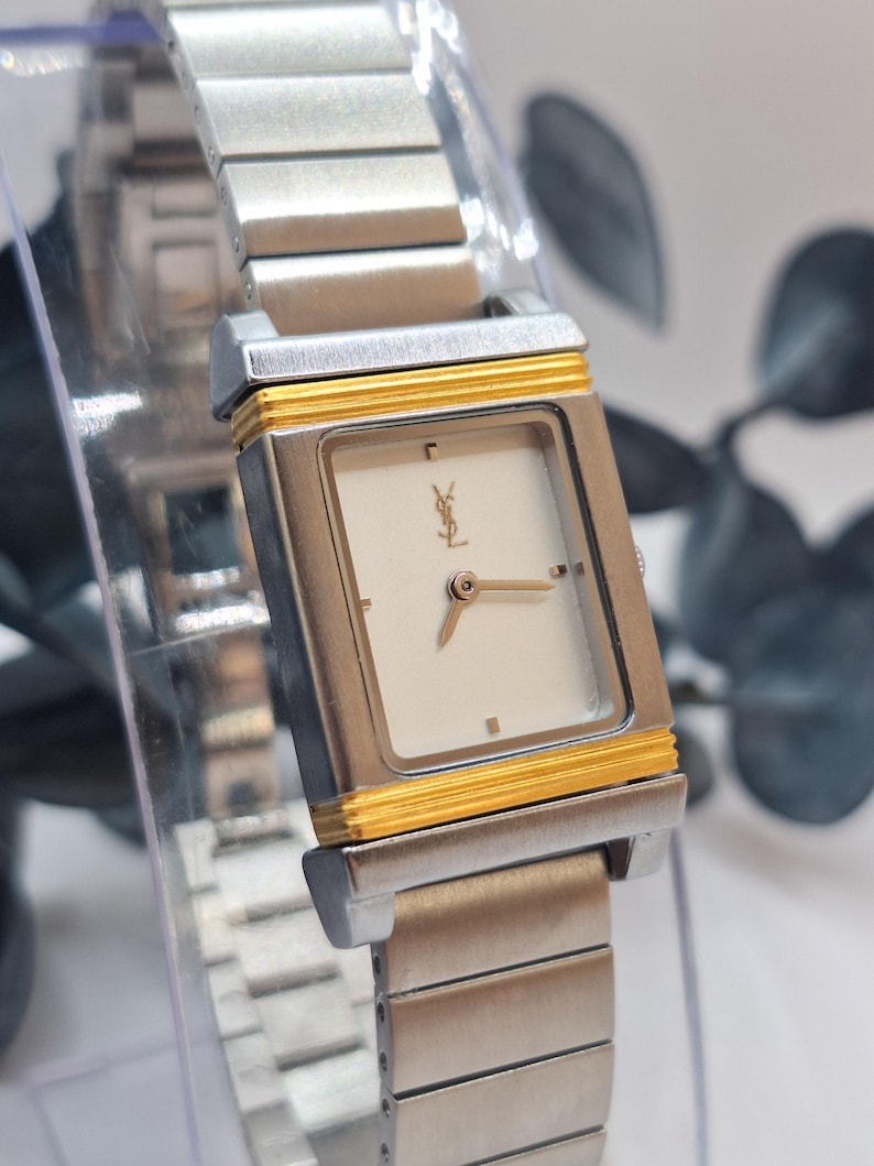 May include: A silver-toned wristwatch with a rectangular face and a gold-colored border. The watch features a white dial with gold hands and a logo. The watch band is made of silver-toned metal links.