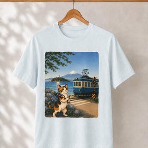 May include: A light blue short-sleeved t-shirt featuring a graphic print of a calico cat carrying a bundle on a stick, standing near a blue train, a railway crossing, purple hydrangea flowers, a body of water, and a snow-capped mountain.