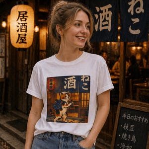 以下が含まれることがあります： 日本のスタイルのイラストが描かれた白いTシャツ。猫がバッグとボトルを持っており、日本語のテキストが書かれています。Tシャツは青いジーンズと組み合わせています。背景には日本語のテキストが書かれた提灯が見えます。