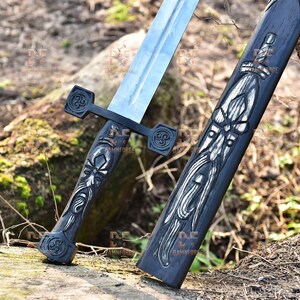 May include: A medieval-style sword with a silver blade and a black handle. The handle and sheath are intricately carved with white and silver designs. The sword is resting on a log in a natural outdoor setting.