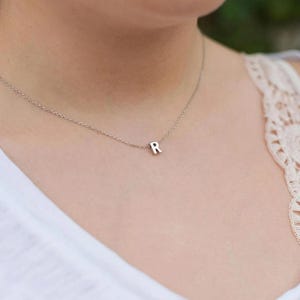 May include: A delicate silver necklace with a small, capital letter "R" pendant. The pendant is centered on a fine chain, creating a minimalist and elegant look. The necklace is shown against a soft background.