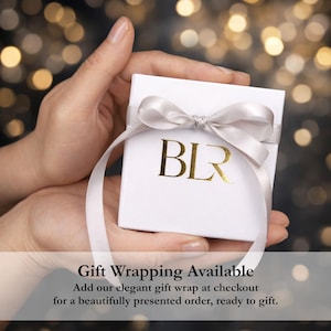 Elegant jewelry gift wrapping shown in a hand, featuring a white presentation box tied with a satin ribbon and gold BLR logo against a softly blurred light background. Gift wrapping is available at checkout for a beautifully presented order ready to give, making this bolo tie necklace, gold lariat necklace or statement jewelry piece a thoughtful gift for her.