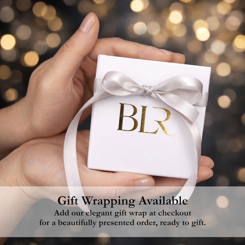 Elegant jewelry gift wrapping shown in a hand, featuring a white presentation box tied with a satin ribbon and gold BLR logo against a softly blurred light background. Gift wrapping is available at checkout for a beautifully presented order ready to give, making this bolo tie necklace, gold lariat necklace or statement jewelry piece a thoughtful gift for her.