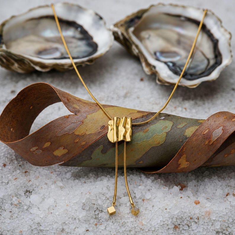 Gold bolo tie necklace displayed across textured natural bark with oyster shells in the background. The long gold lariat necklace features a polished sculptural wave slider, fine snake chain and two tassel drops with small square charms. Artistic styling highlights the organic statement necklace design, warm gold finish and distinctive adjustable Y necklace shape for modern everyday jewelry.