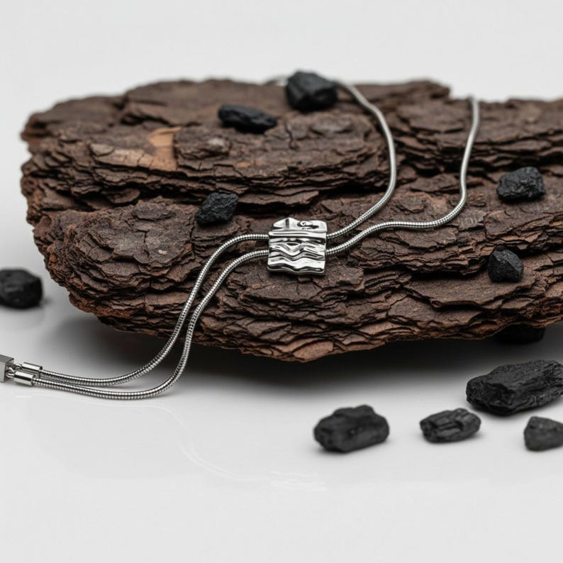 Silver bolo tie lariat necklace styled across textured dark tree bark with scattered black stones. The polished sculptural slider contrasts with the natural surface while the long snake chain forms an adjustable Y necklace with two tassel drops. Artistic product photography highlights the organic statement necklace design, silver tone finish and modern long necklace silhouette.
