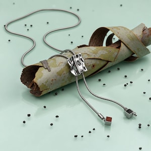 Silver bolo tie lariat necklace displayed on pale green eucalyptus bark with small black stones scattered across the background. The polished sculptural slider creates an organic focal point while the long snake chain forms an adjustable Y necklace with two tassel drops. Creative product styling highlights the contemporary statement necklace design and sleek silver tone finish.