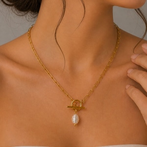 May include: Gold-toned necklace featuring a toggle clasp and a dangling pearl pendant. The chain has a rectangular link design. The pearl is white with a slight luster. The necklace is worn around the neck.