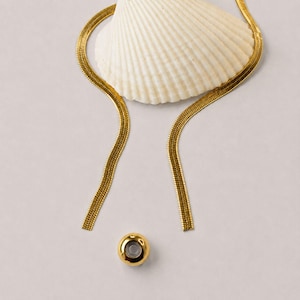 Adjustable gold snake chain lariat necklace with removable slider bead displayed beside a white seashell. Modern bolo tie necklace featuring a polished gold slider and sleek flat snake chain. Waterproof jewelry designed for versatile styling as a Y necklace, long necklace, minimalist necklace, or layering necklace. Elegant gift for her with contemporary coastal-inspired design and adjustable fit.