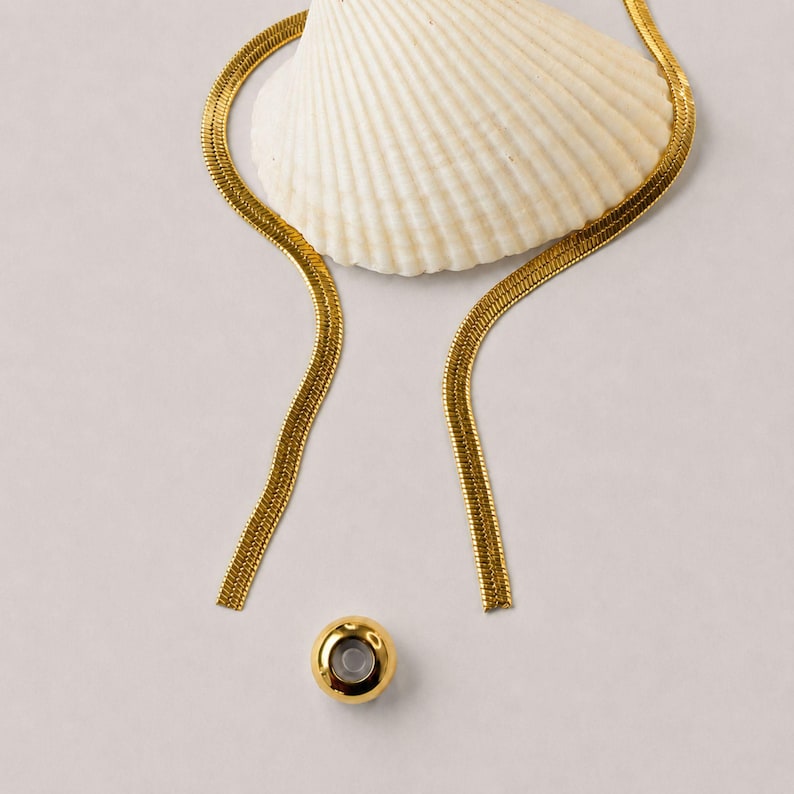 Adjustable gold snake chain lariat necklace with removable slider bead displayed beside a white seashell. Modern bolo tie necklace featuring a polished gold slider and sleek flat snake chain. Waterproof jewelry designed for versatile styling as a Y necklace, long necklace, minimalist necklace, or layering necklace. Elegant gift for her with contemporary coastal-inspired design and adjustable fit.