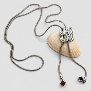 Silver tone bolo tie necklace arranged around a natural seashell on a clean light background. The long snake chain passes through a polished sculptural wavy slider and extends into two tassel drops finished with geometric square charms. Product styling highlights the adjustable lariat necklace construction, organic statement design and contemporary Y necklace silhouette.