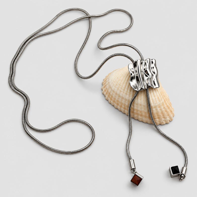 Silver tone bolo tie necklace arranged around a natural seashell on a clean light background. The long snake chain passes through a polished sculptural wavy slider and extends into two tassel drops finished with geometric square charms. Product styling highlights the adjustable lariat necklace construction, organic statement design and contemporary Y necklace silhouette.