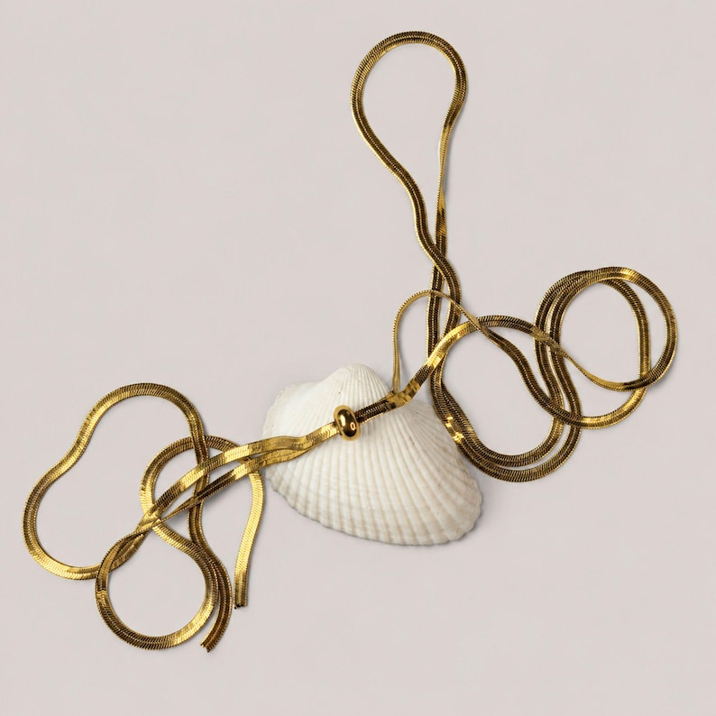 Adjustable gold snake chain necklace arranged artistically around a white seashell. Modern bolo tie necklace with removable slider bead and flexible styling options. Waterproof jewelry suitable as a long necklace, Y necklace, layering necklace, and everyday accessory. Minimalist gold jewelry inspired by clean lines and effortless elegance.