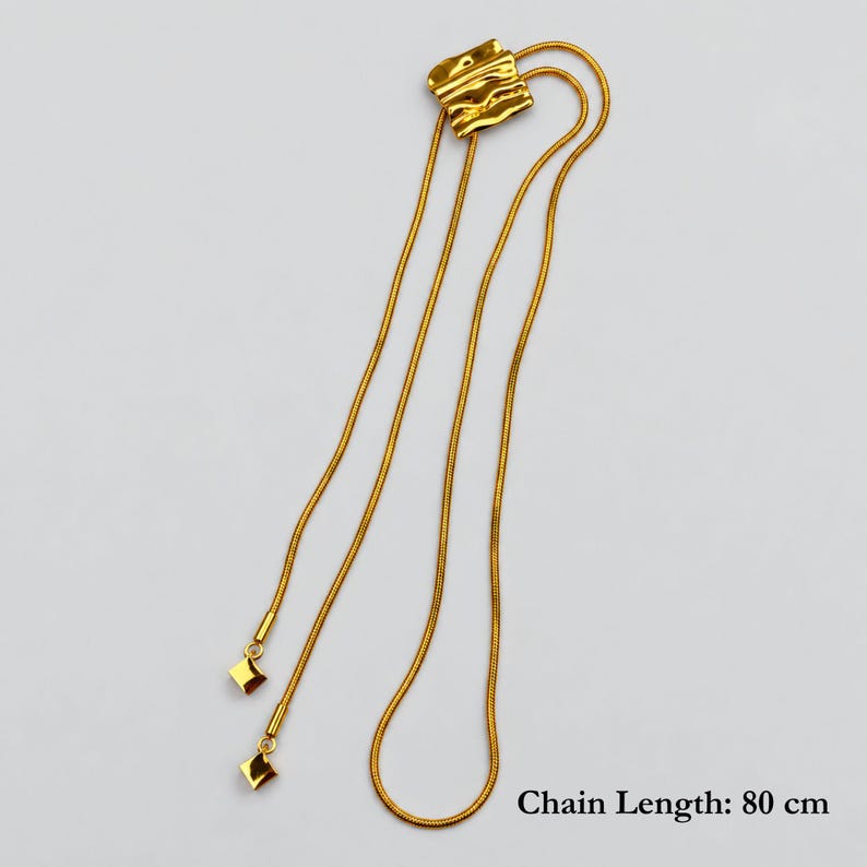 Full-length product view of a gold bolo tie lariat necklace arranged on a light neutral background, showing the complete 80 cm snake chain and two tassel ends with geometric square charms. The sculptural wavy slider creates the adjustable Y necklace shape. Clear flat lay demonstrates the generous length and versatile construction of this long gold statement necklace.