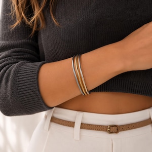 May include: Three metal bangles in silver and gold tones. The bracelets are stacked on an arm, with a dark grey sweater in the background. The bangles are simple and elegant, suitable for everyday wear.