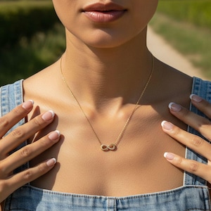 May include: A gold necklace featuring an infinity symbol pendant. The necklace is displayed on a person's chest, with hands and a denim top in view. The infinity symbol is a popular design in jewellery.