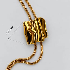 Macro close-up of the gold bolo tie necklace slider showing its sculptural wavy polished surface and approximately 20 mm height. Detailed product view highlights the reflective gold finish, snake chain passing through the statement slider and organic textured design. A close look at the central feature of this adjustable gold lariat necklace, Y necklace and long statement jewelry piece.