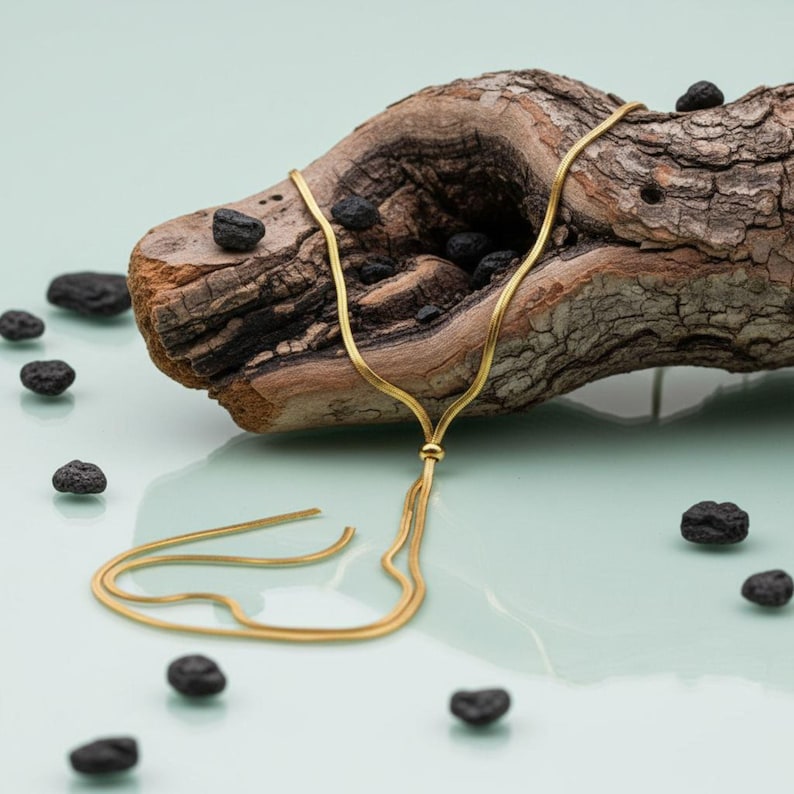 Gold lariat necklace displayed on textured driftwood with decorative black stones. Adjustable snake chain necklace featuring a polished slider bead and long flowing chain ends. Waterproof jewelry designed as a modern Y necklace, layering necklace, and statement accessory. Elegant gift for her with minimalist and contemporary appeal.