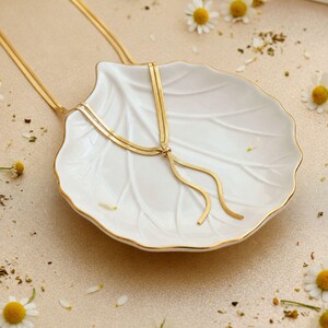 Elegant gold snake chain lariat necklace displayed on white ceramic shell dish with soft floral accents, highlighting adjustable slider bead and flowing tassel ends. Minimalist bolo tie necklace for women, waterproof jewelry, modern gold Y necklace, refined gift idea, stylish long necklace, perfect for feminine everyday wear or special occasions.