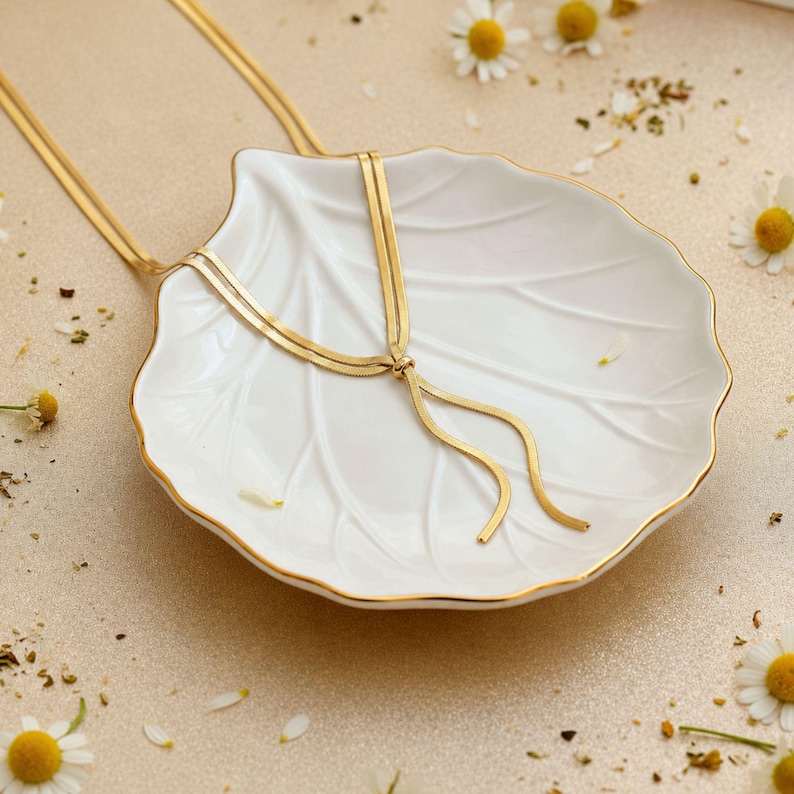 Elegant gold snake chain lariat necklace displayed on white ceramic shell dish with soft floral accents, highlighting adjustable slider bead and flowing tassel ends. Minimalist bolo tie necklace for women, waterproof jewelry, modern gold Y necklace, refined gift idea, stylish long necklace, perfect for feminine everyday wear or special occasions.
