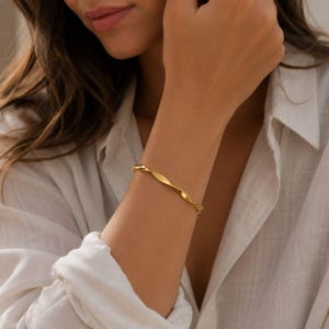 May include: A gold-toned bangle bracelet featuring a continuous twisted, spiral design. The metal surface has a smooth, polished finish that reflects light along the curved edges of the band, creating a simple and uniform circular shape.
