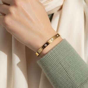 May include: A gold bangle bracelet featuring several small, round, clear gemstones set into the band. The bracelet is worn on a wrist, with a green jumper and cream-coloured fabric in the background.
