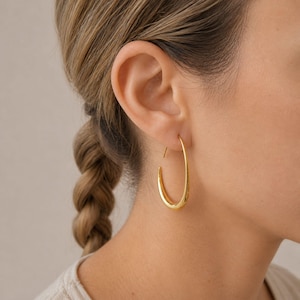 May include: A close-up of a gold-coloured hoop earring. The earring is a simple, elongated oval shape. The earring is worn on the ear of a person with light skin and blonde hair in a braid.