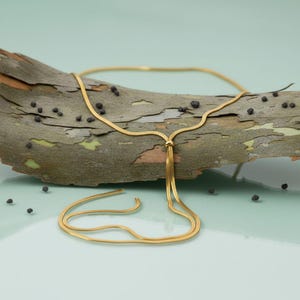 Gold adjustable bolo tie necklace styled on natural eucalyptus bark with soft green background and reflective surface. Waterproof snake chain necklace featuring a polished slider bead and elegant drop chains. Minimalist lariat necklace with modern design, ideal for layering, everyday wear, gifting, and sophisticated coastal-inspired styling.