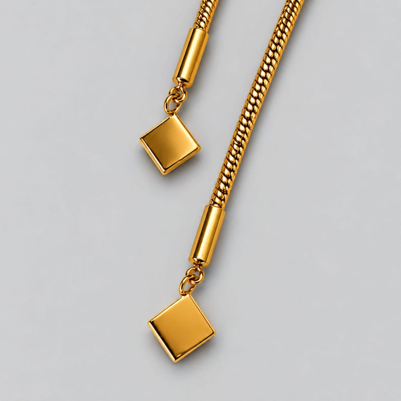 Macro detail of the two tassel ends on a gold bolo tie lariat necklace, showing the flexible snake chain, polished cylindrical end caps, connecting rings and geometric square charms. Close-up product photography highlights the smooth reflective gold finish and fine construction details of this long Y necklace, statement lariat necklace and adjustable slider jewelry design.