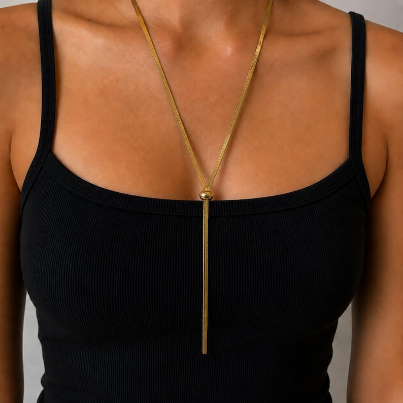 Front close-up of gold minimalist lariat necklace on model wearing black ribbed camisole, featuring sleek snake chain, polished adjustable bead, and long Y tassel drop. Waterproof women’s jewelry, modern bolo necklace, elegant layering piece, stylish long necklace, refined statement accessory, gift for her with versatile everyday appeal.